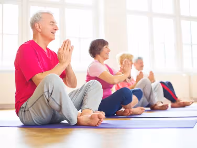 A group of older adults sit cross-legged on yoga mats indoors, with hands pressed together in a prayer position, smiling and participating in a yoga or meditation class.