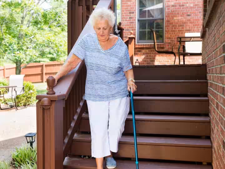 An older woman with white hair descends outdoor wooden stairs using a cane, holding the railing for support. She wears a striped shirt, white pants, and blue shoes. A brick house and patio are visible in the background.