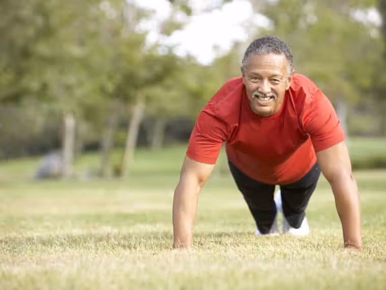 A man wearing a red shirt and black pants does a push-up on grass in a park, smiling at the camera with trees in the blurred background.