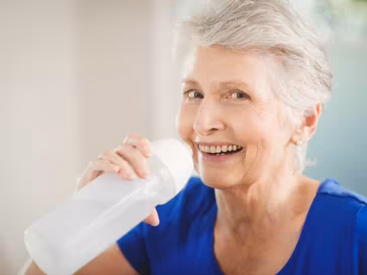 Smiling older woman with short gray hair wearing a blue shirt, holding a water bottle and appearing to take a drink indoors.
