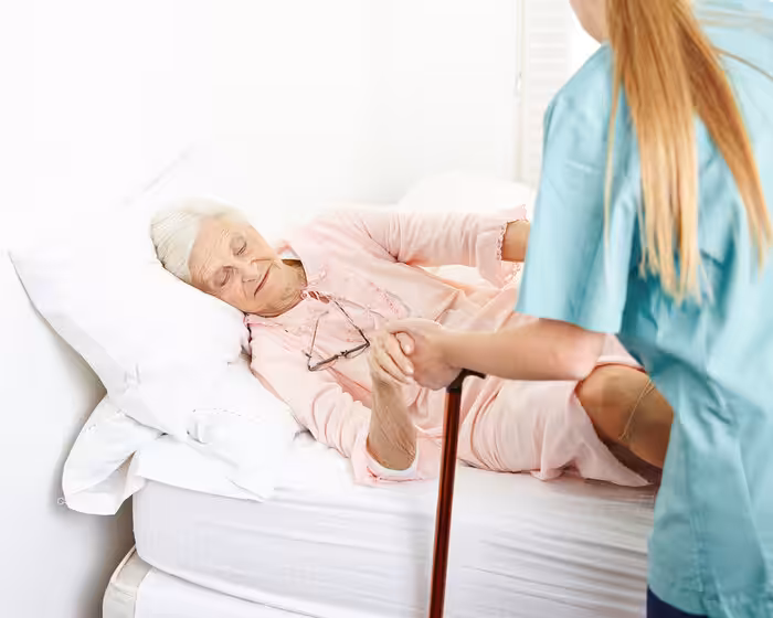 An elderly woman in pink pajamas lies in bed, holding a cane and the hand of a caregiver wearing scrubs, who is helping her sit up. The setting appears to be a bright, clean bedroom.