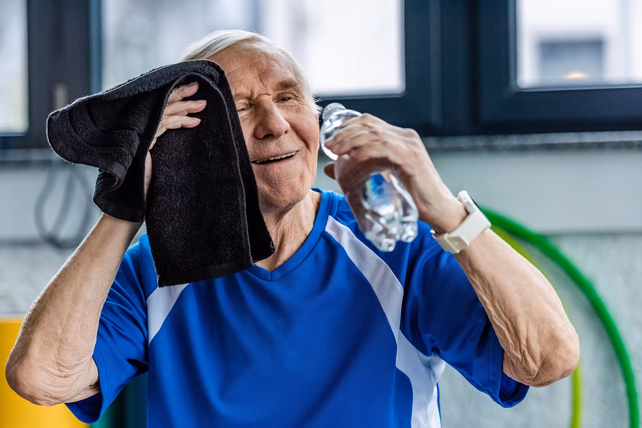 An older man in a blue athletic shirt wipes his face with a black towel and holds a water bottle, smiling and looking relaxed after exercising indoors.
