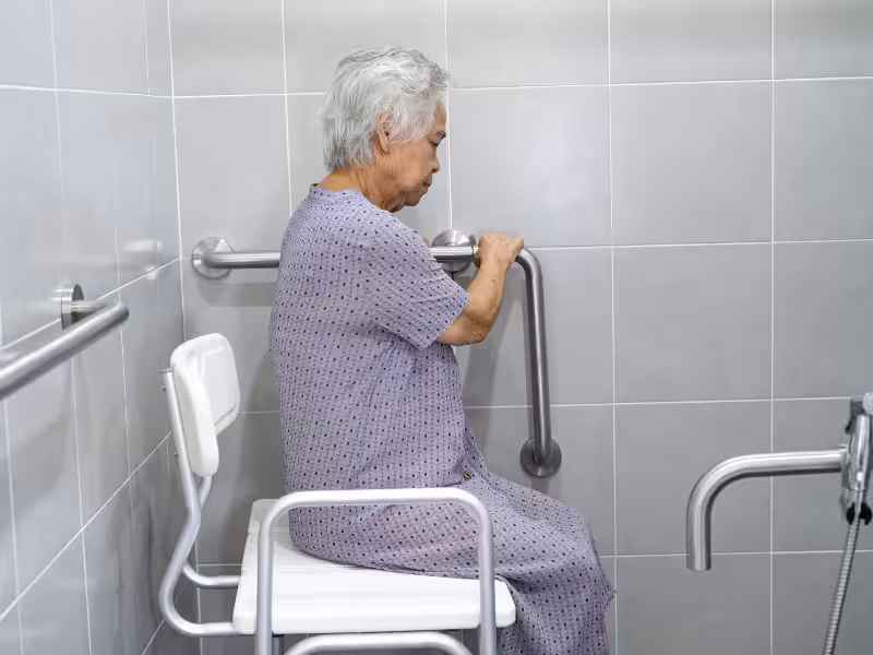 An elderly person with gray hair in a hospital gown sits on a shower chair, holding onto grab bars in a tiled bathroom, indicating an accessible, senior-friendly bathing area.