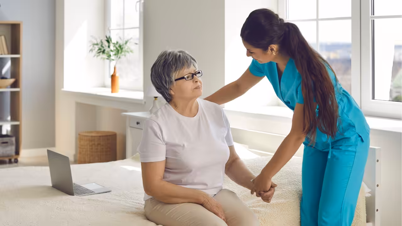 A nurse in blue scrubs helps an older woman with gray hair and glasses, who is sitting on a bed. They are holding hands in a bright room with large windows and a laptop on the bed.