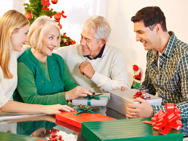 Four people sit around a table exchanging wrapped gifts, smiling and enjoying each others company. A decorated Christmas tree is visible in the background, adding a festive touch to the scene.