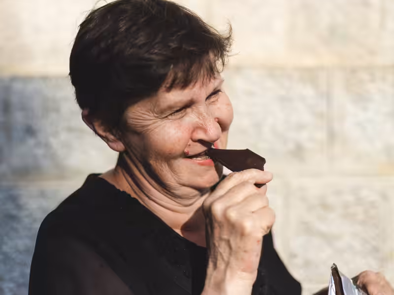 An older woman with short brown hair smiles as she prepares to take a bite of a piece of chocolate, standing in front of a light-colored textured wall.