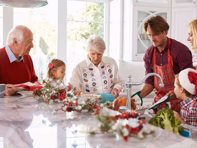 Three generations of a family gather in a bright kitchen, preparing a holiday meal together. The counter is decorated with festive greenery and ornaments, and everyone appears joyful and engaged in cooking.