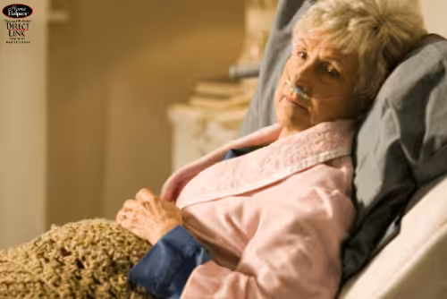 An elderly woman with gray hair lies in bed, wearing a pink robe and an oxygen tube. She looks thoughtful and slightly tired, covered with a knitted blanket.