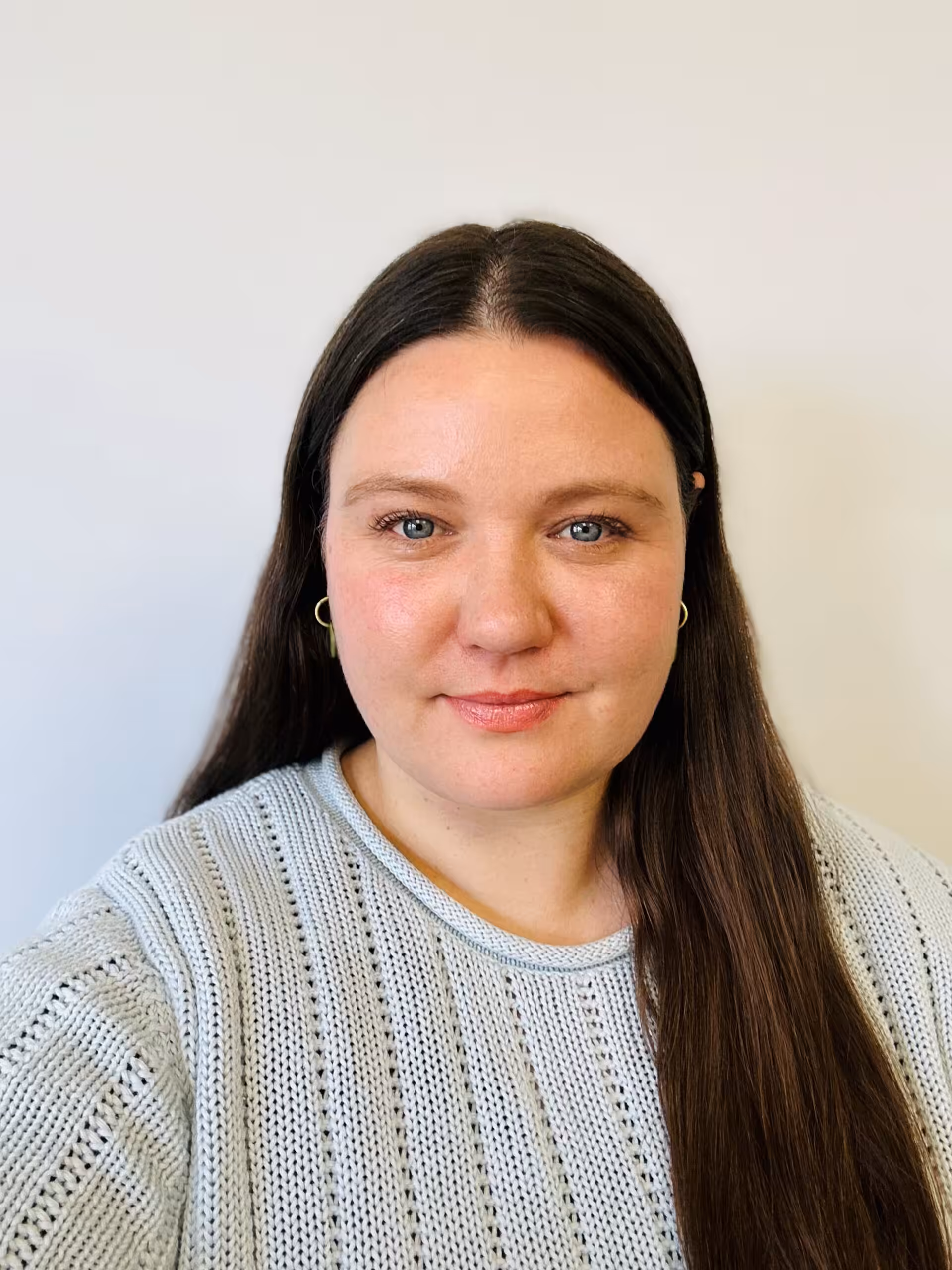 A woman with long brown hair and light skin is wearing a light gray knit sweater and small hoop earrings. She is smiling softly and is set against a plain, light background.