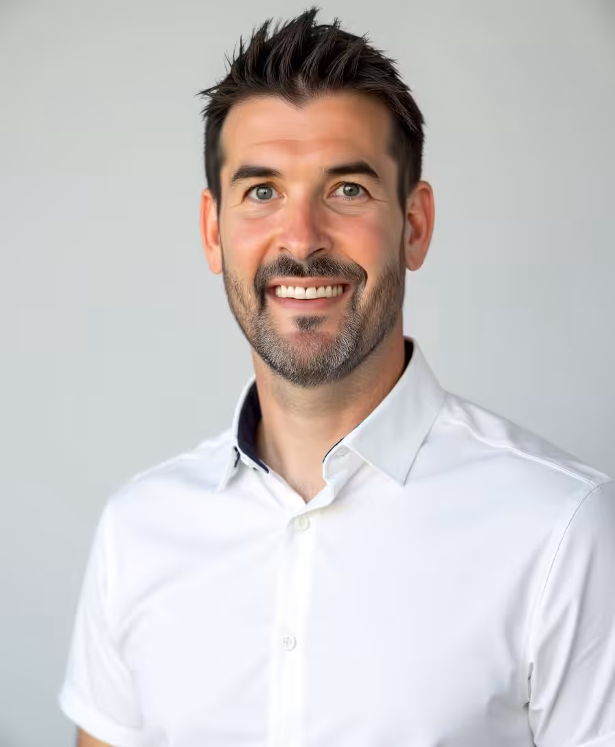 A smiling man with short dark hair and a trimmed beard wearing a white collared shirt, standing against a plain light gray background.