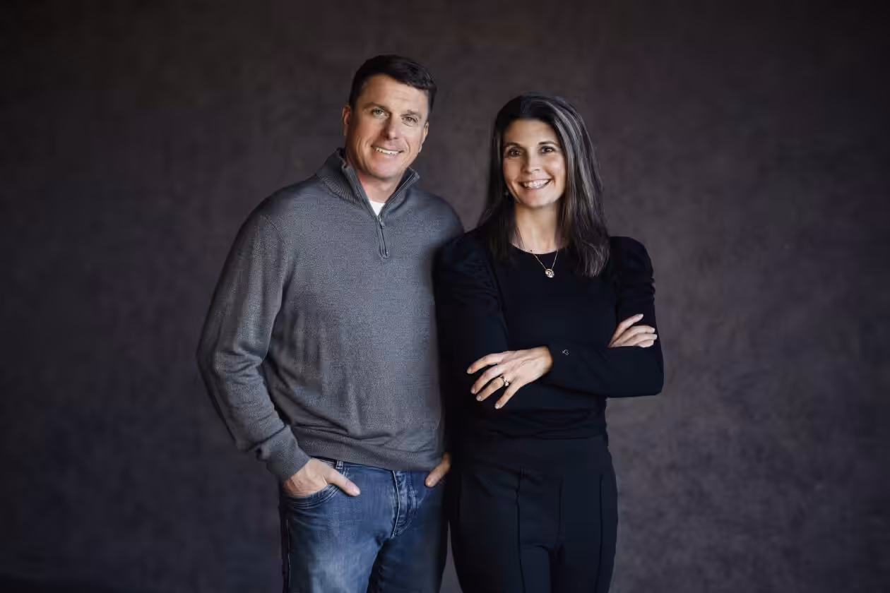 A man in a gray sweater and jeans stands next to a woman in a black shirt and pants. Both are smiling, with the woman’s arms crossed and the man’s hands in his pockets, against a plain dark background.