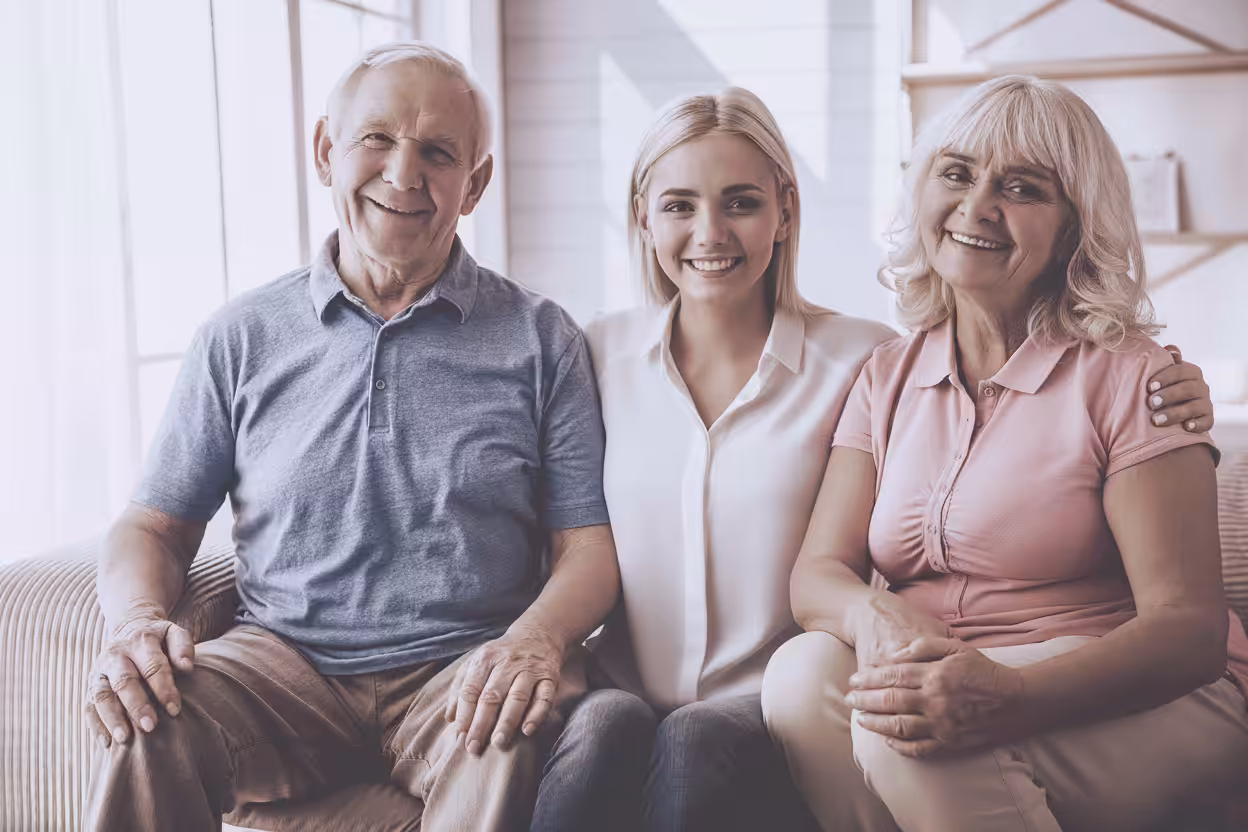 Three smiling people, two older adults and a younger adult, sit close together on a couch in a bright room, showing a warm and happy family moment.