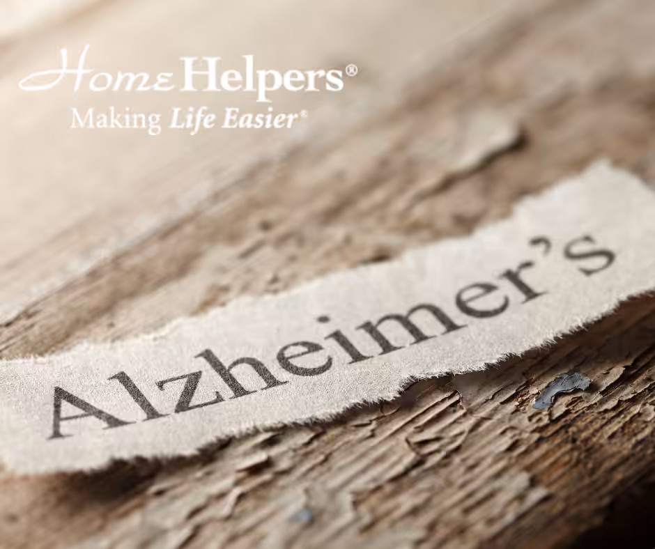 A torn piece of paper with the word “Alzheimer’s” rests on a wooden surface. In the top left corner, the Home Helpers logo and the phrase “Making Life Easier” appear in white text.
