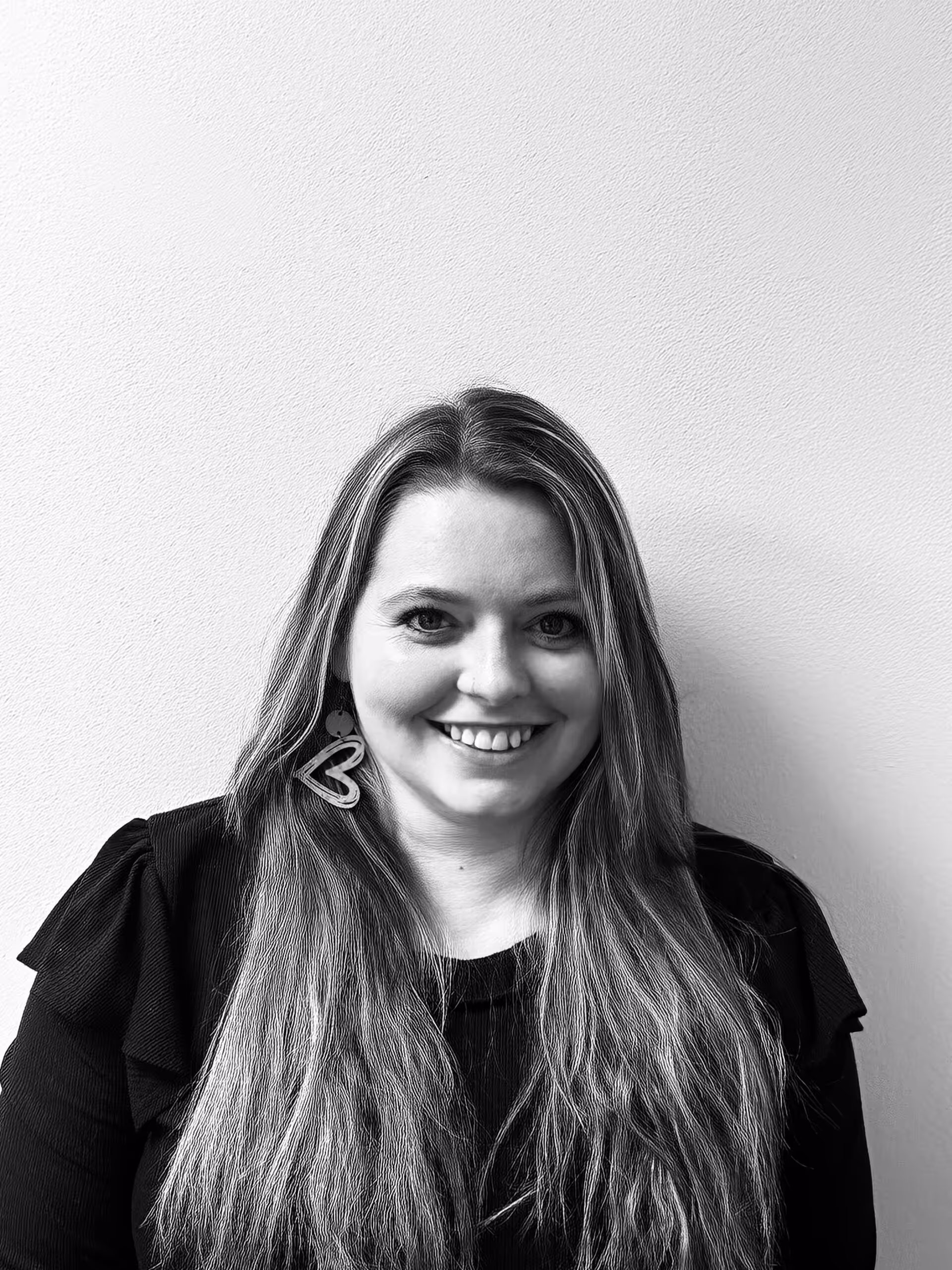 A woman with long, light hair wearing a dark top and large heart-shaped earrings smiles in front of a plain, light-colored wall. The photo is in black and white.