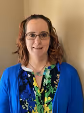 A woman with shoulder-length wavy brown hair, glasses, and a blue cardigan over a colorful blouse stands indoors against a beige wall, smiling at the camera.