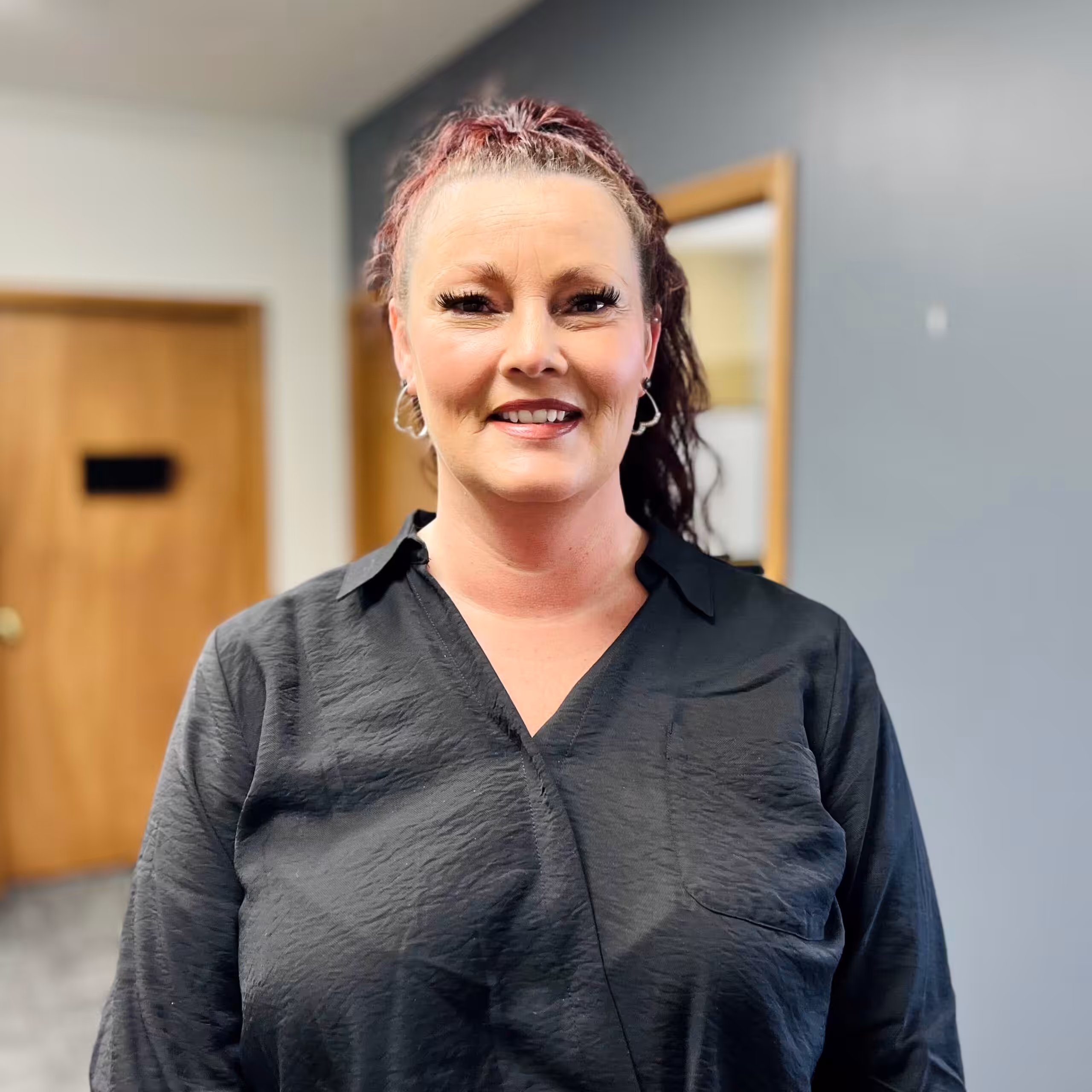 A woman with curly red hair in a ponytail, wearing a black textured blouse, stands indoors in front of wooden doors and a gray wall, smiling at the camera.