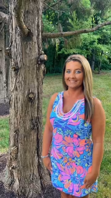 A woman wearing a colorful, floral sleeveless dress stands outside next to a tree, smiling at the camera with green grass and lush trees in the background.