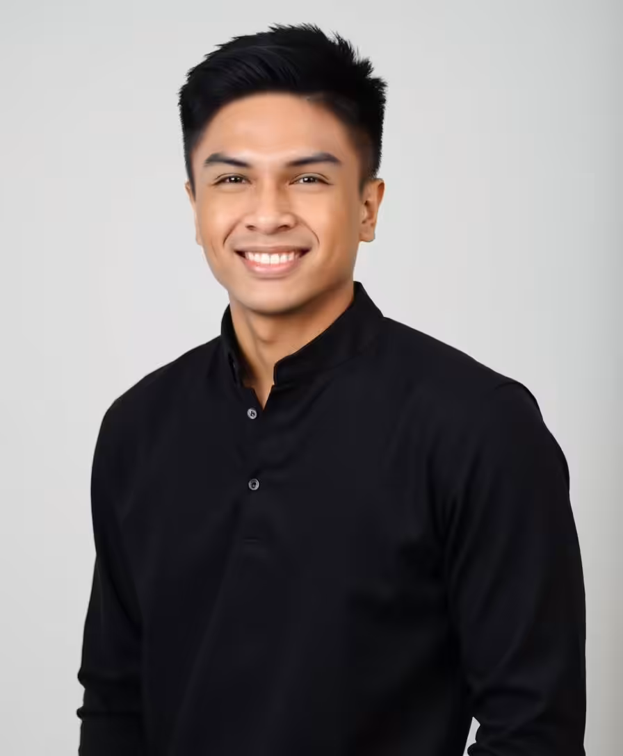 A young man with short black hair smiles at the camera. He is wearing a black button-up shirt with a stand-up collar, and the background is plain and light-colored.