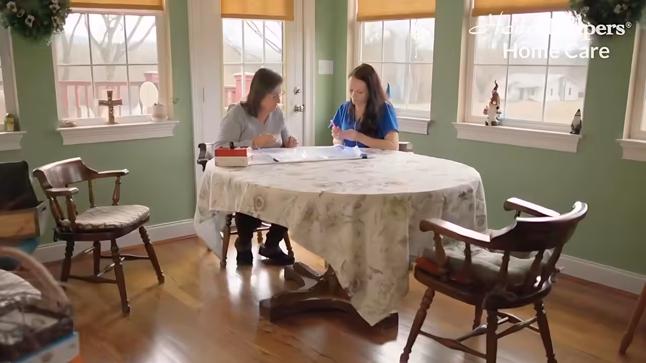 Two women sit at a round dining table in a bright, homey room. One woman wears blue scrubs and writes on a clipboard, while the other observes. Text in the corner reads Home Helpers Home Care.