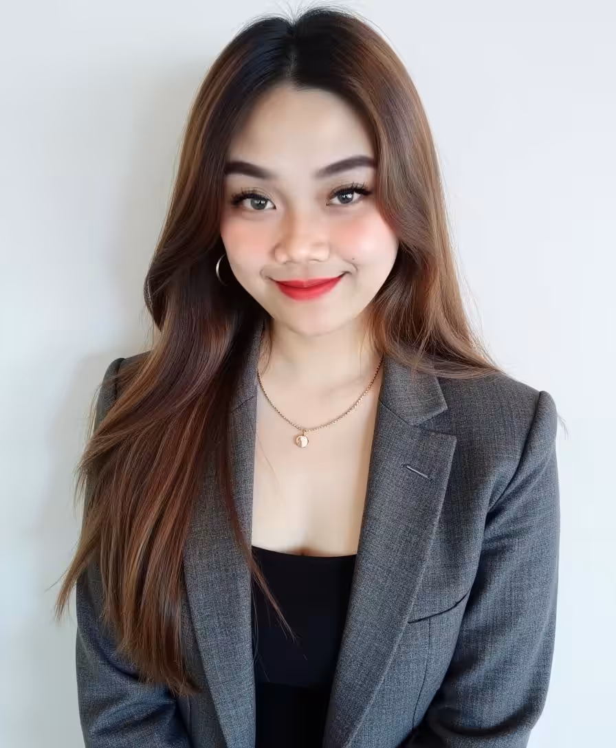 A young woman with long brown hair, wearing a gray blazer over a black top, gold hoop earrings, and a necklace, smiles while standing in front of a plain white background.