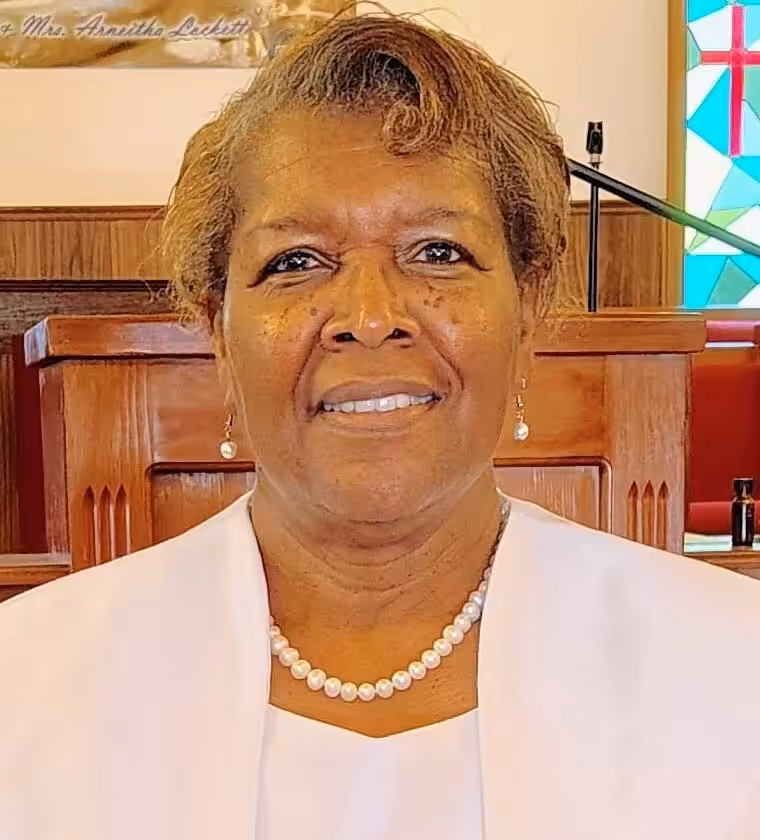A woman with short, curly hair wearing a white outfit and a pearl necklace stands smiling in a church with wooden pews and colorful stained glass in the background.