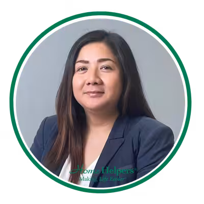 A woman with long, straight dark hair wearing a navy blazer and light top poses against a gray background, framed by a green circular border with Home Helpers logo at the bottom.