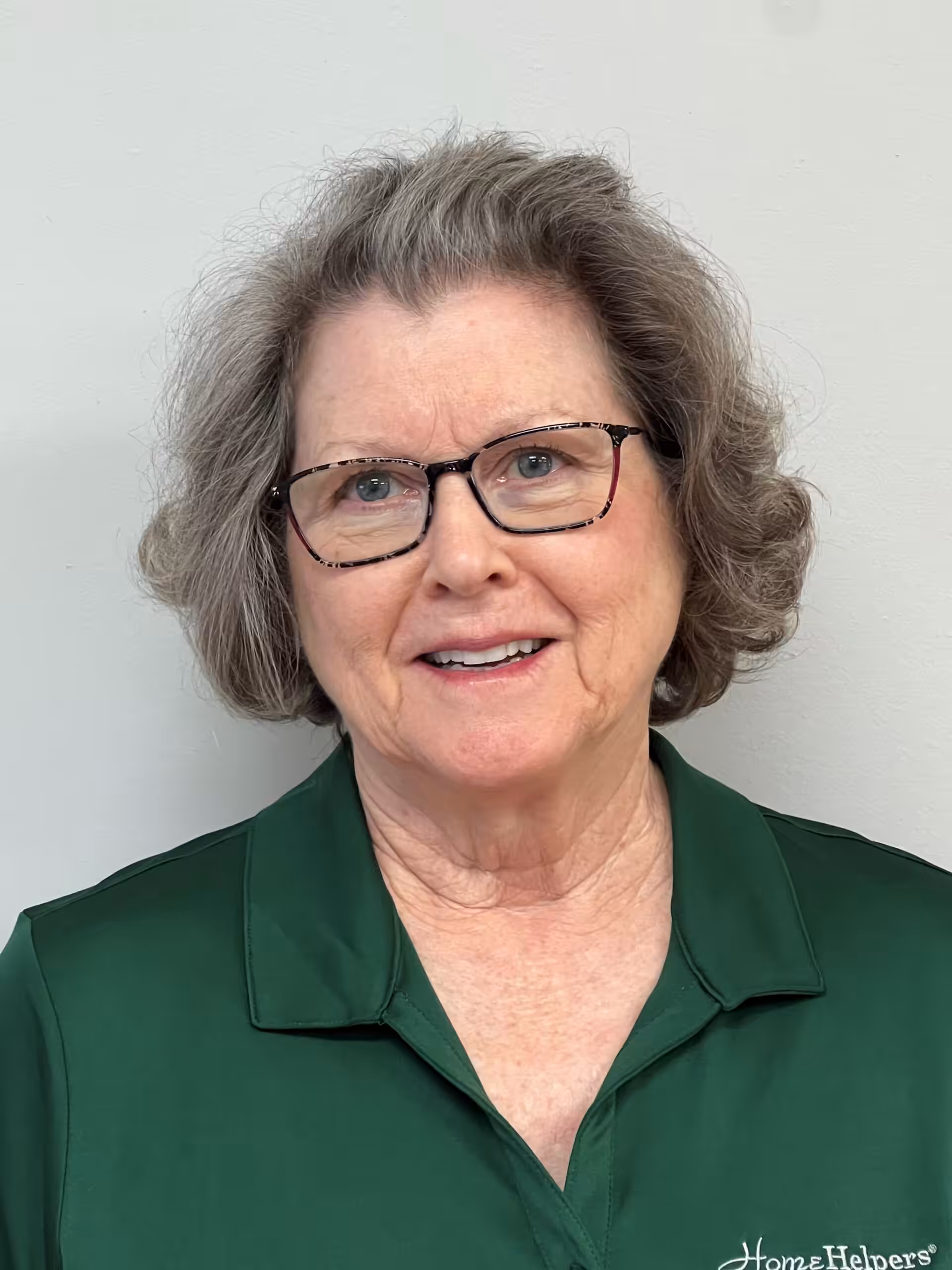 An older woman with short, curly gray hair and glasses is smiling at the camera. She is wearing a dark green collared shirt with a Home Helpers logo on the chest, standing in front of a plain light-colored wall.