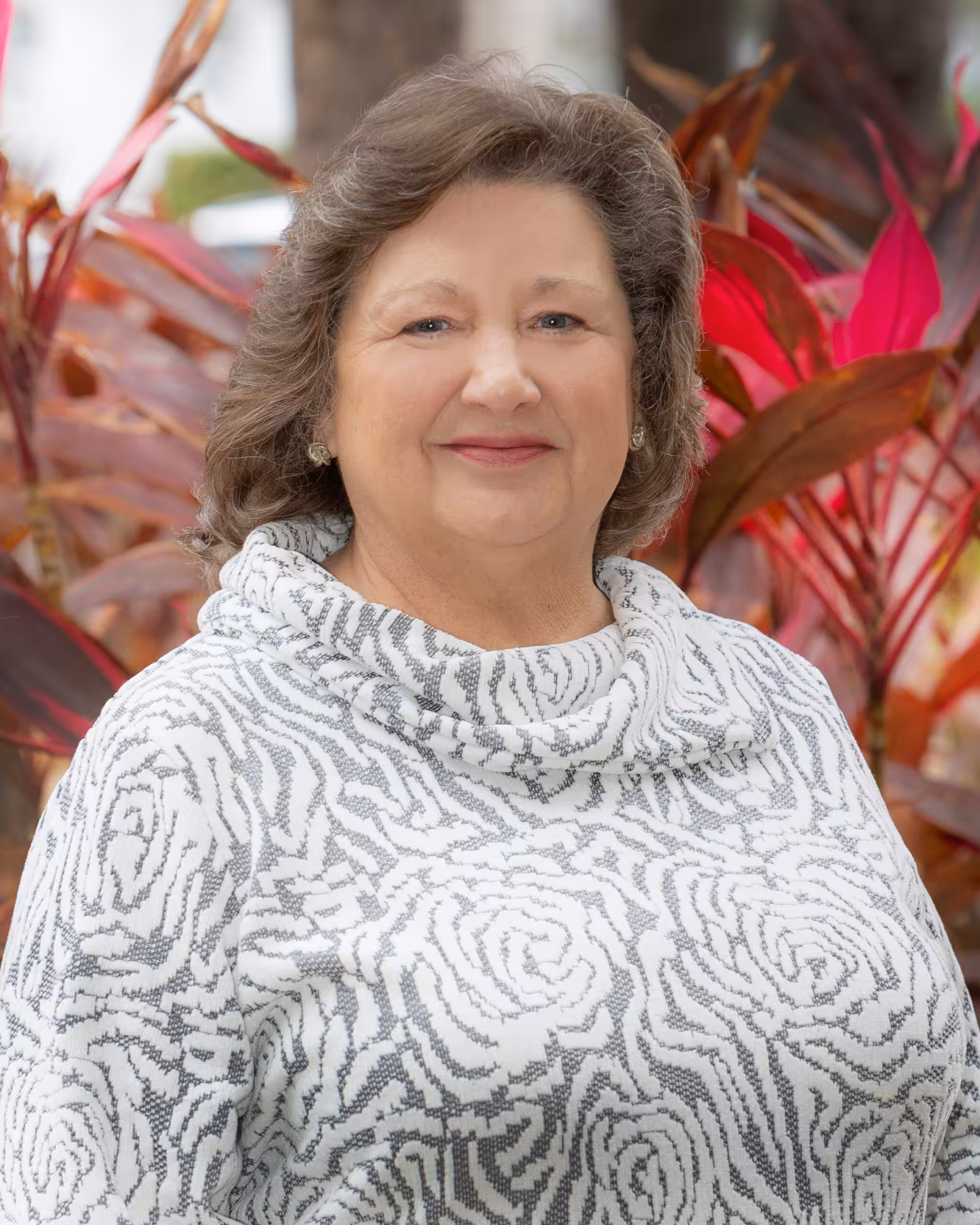 A woman with wavy brown hair smiles while standing in front of red and green tropical plants. She is wearing a white and gray patterned sweater with a cowl neck.