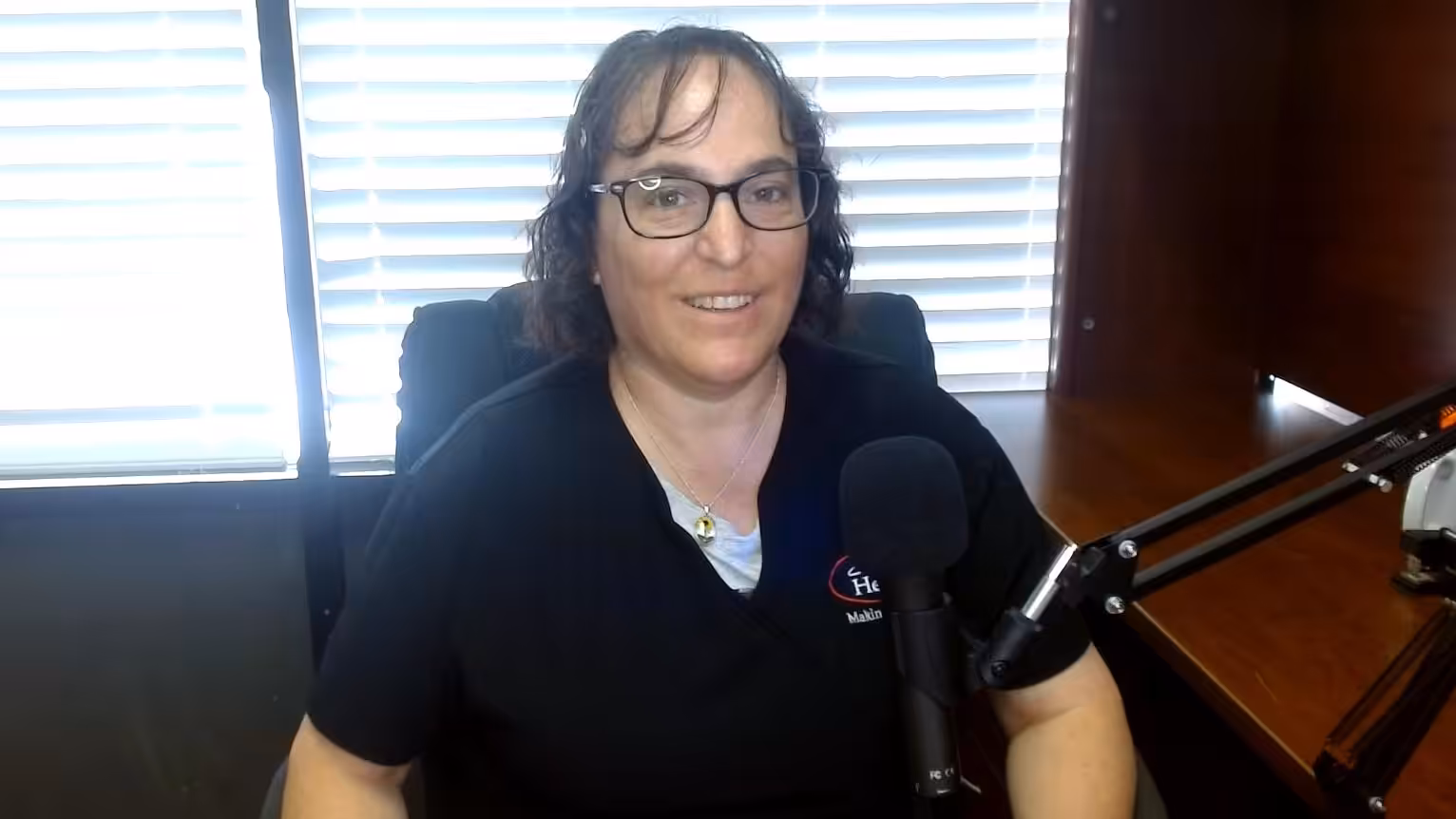 A woman with glasses and dark, wavy hair is smiling while sitting at a desk with a microphone in front of her. She is wearing a black shirt, and there are window blinds and office shelves in the background.