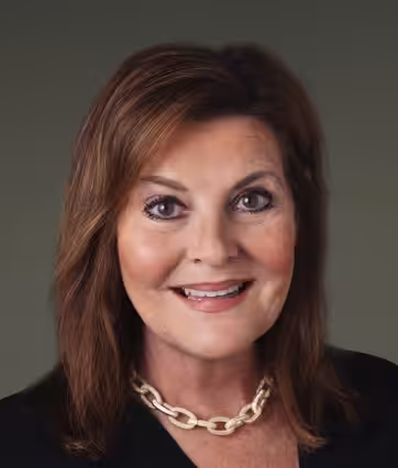 A woman with shoulder-length brown hair smiles at the camera. She is wearing a black top and a chunky gold chain necklace, with a neutral green background.