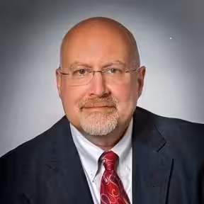 A middle-aged man with glasses, a bald head, and a trimmed gray beard wears a dark suit, white shirt, and red patterned tie, posing against a plain gray background.