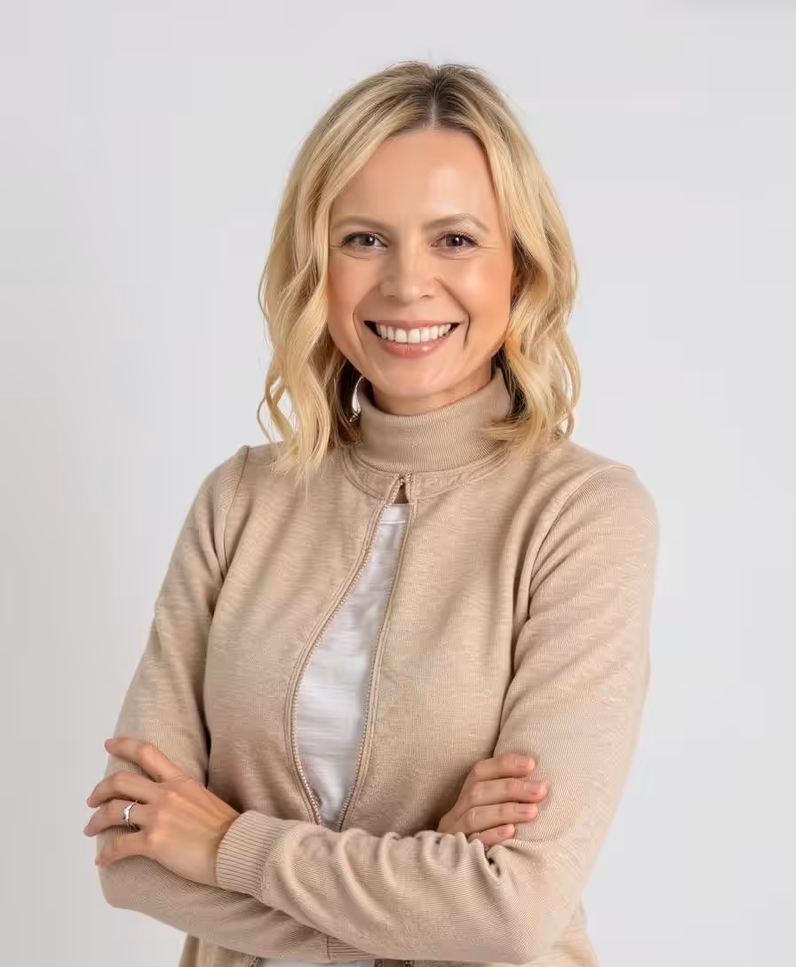 A smiling woman with blonde hair in loose waves wearing a beige turtleneck sweater and a white shirt stands with arms crossed against a plain light background.