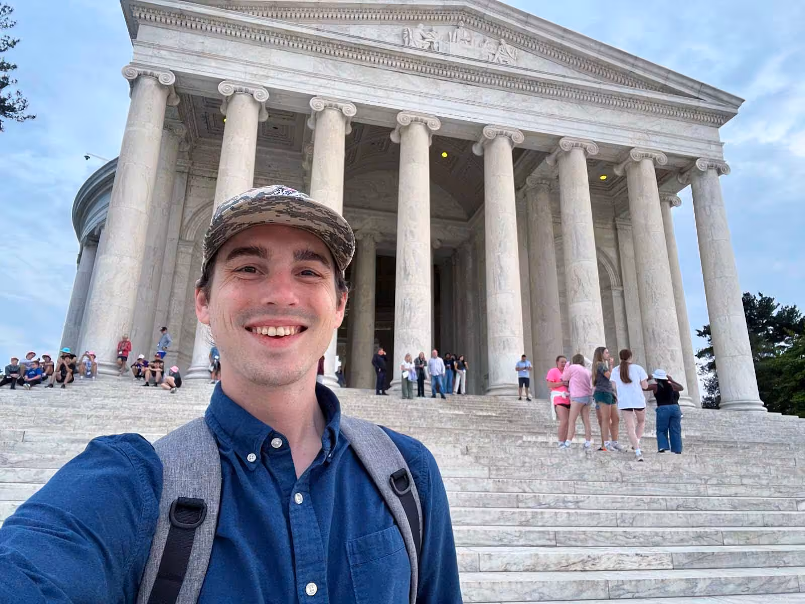 A smiling person wearing a cap and backpack takes a selfie on the marble steps of a neoclassical building with tall columns. Groups of people are scattered on the steps and gathered near the entrance.