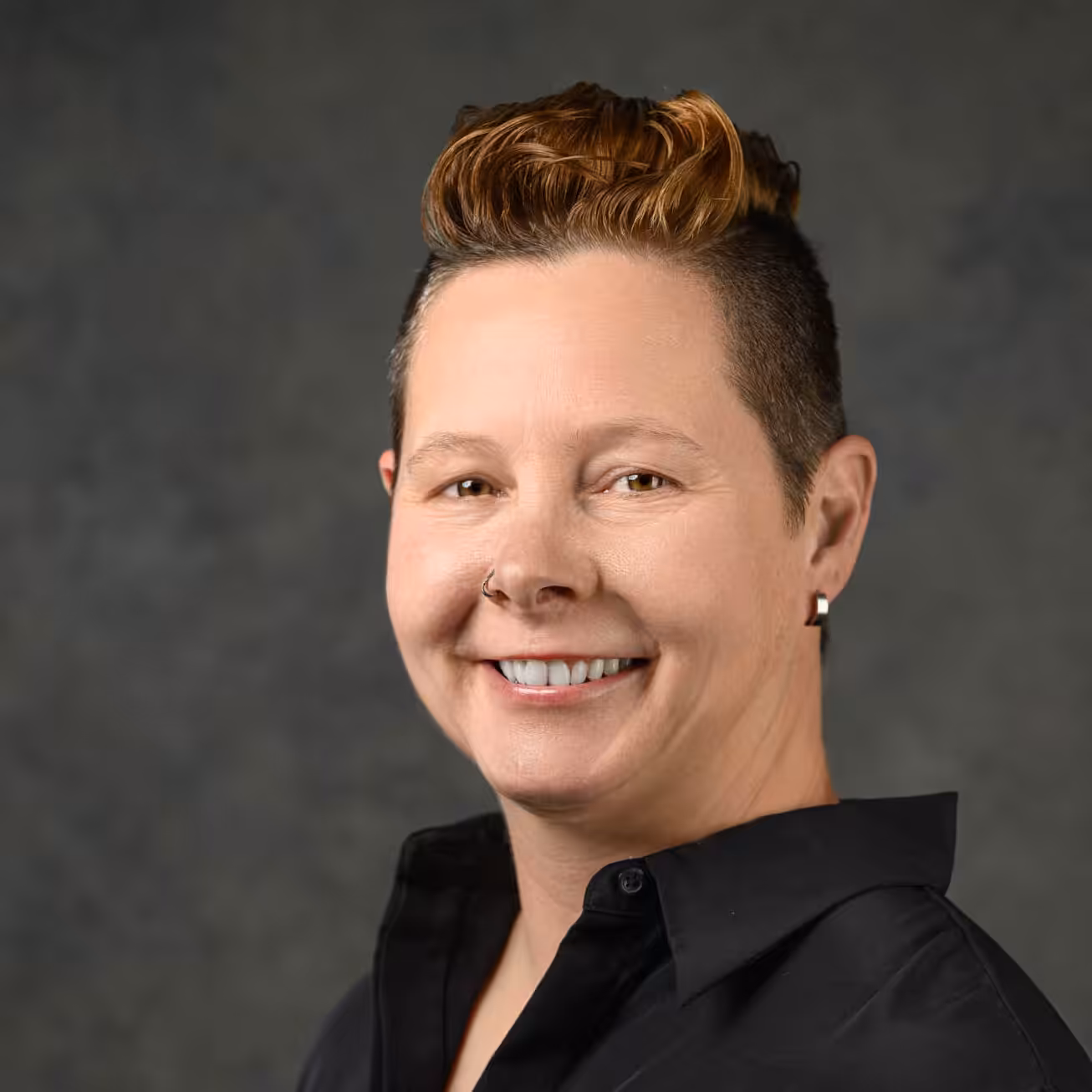 A person with short, styled brown hair, wearing a black collared shirt and a small earring, smiles at the camera in front of a gray backdrop.