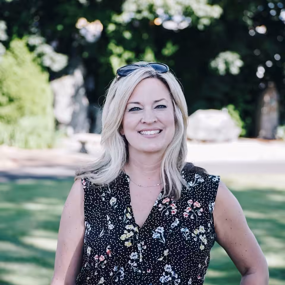 A woman with blonde hair, wearing sunglasses on her head and a sleeveless floral top, smiles while standing outdoors in a sunny, green park with trees in the background.