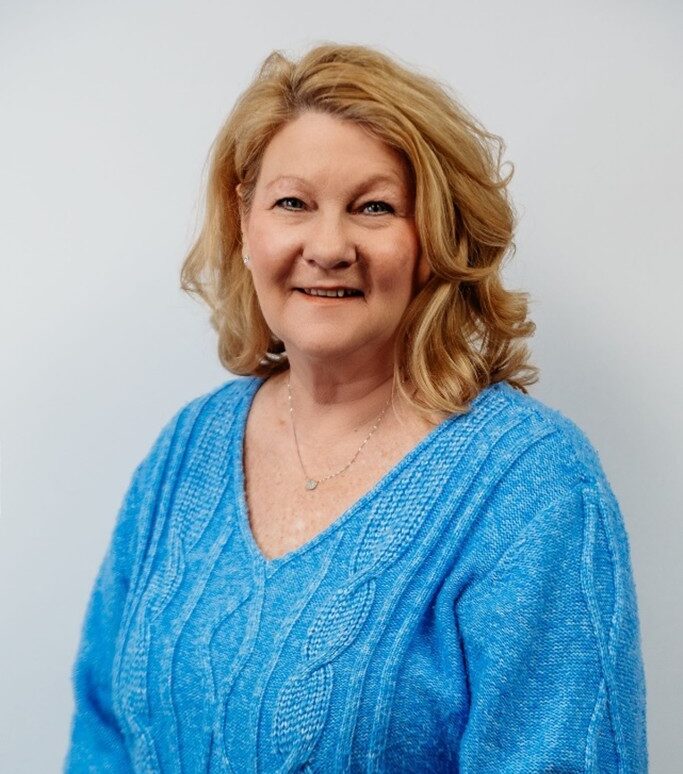 A woman with shoulder-length blonde hair smiles at the camera. She is wearing a bright blue textured sweater and a delicate necklace, standing in front of a plain light background.