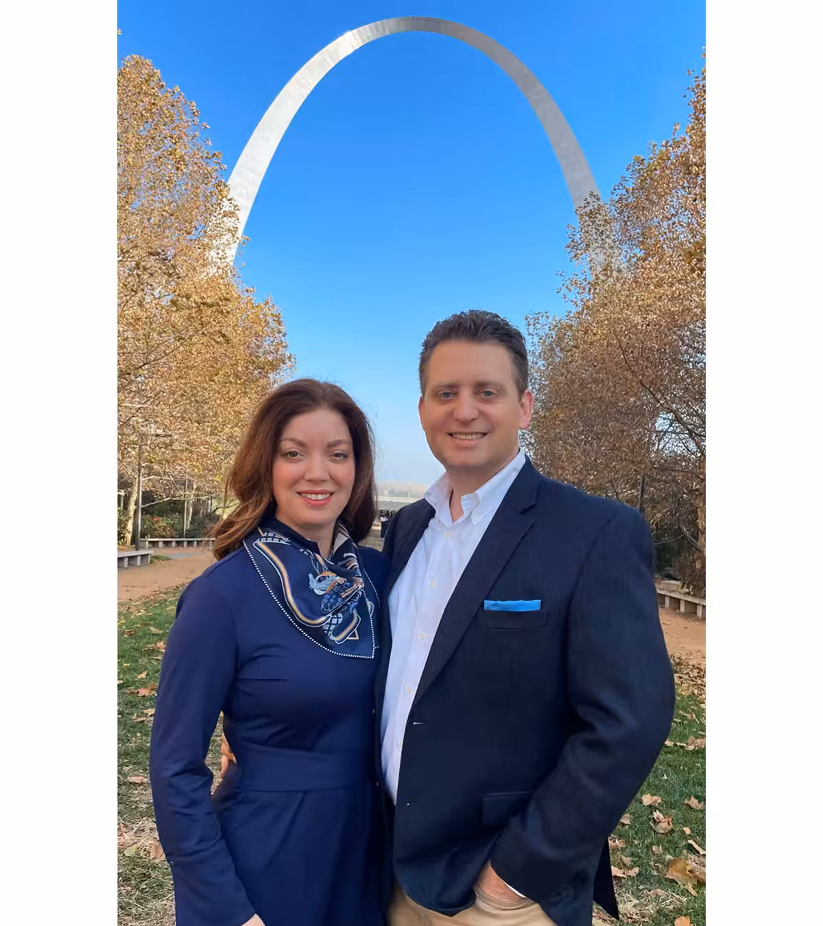 A smiling couple stands outdoors in front of the Gateway Arch in St. Louis, surrounded by autumn trees and a clear blue sky. The woman wears a navy dress and scarf; the man wears a dark blazer and white shirt.