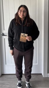 A person with long dark hair and glasses stands indoors, wearing a black hoodie, dark pants, and black-and-white sneakers, holding a gold Visa gift card in their right hand and smiling slightly.