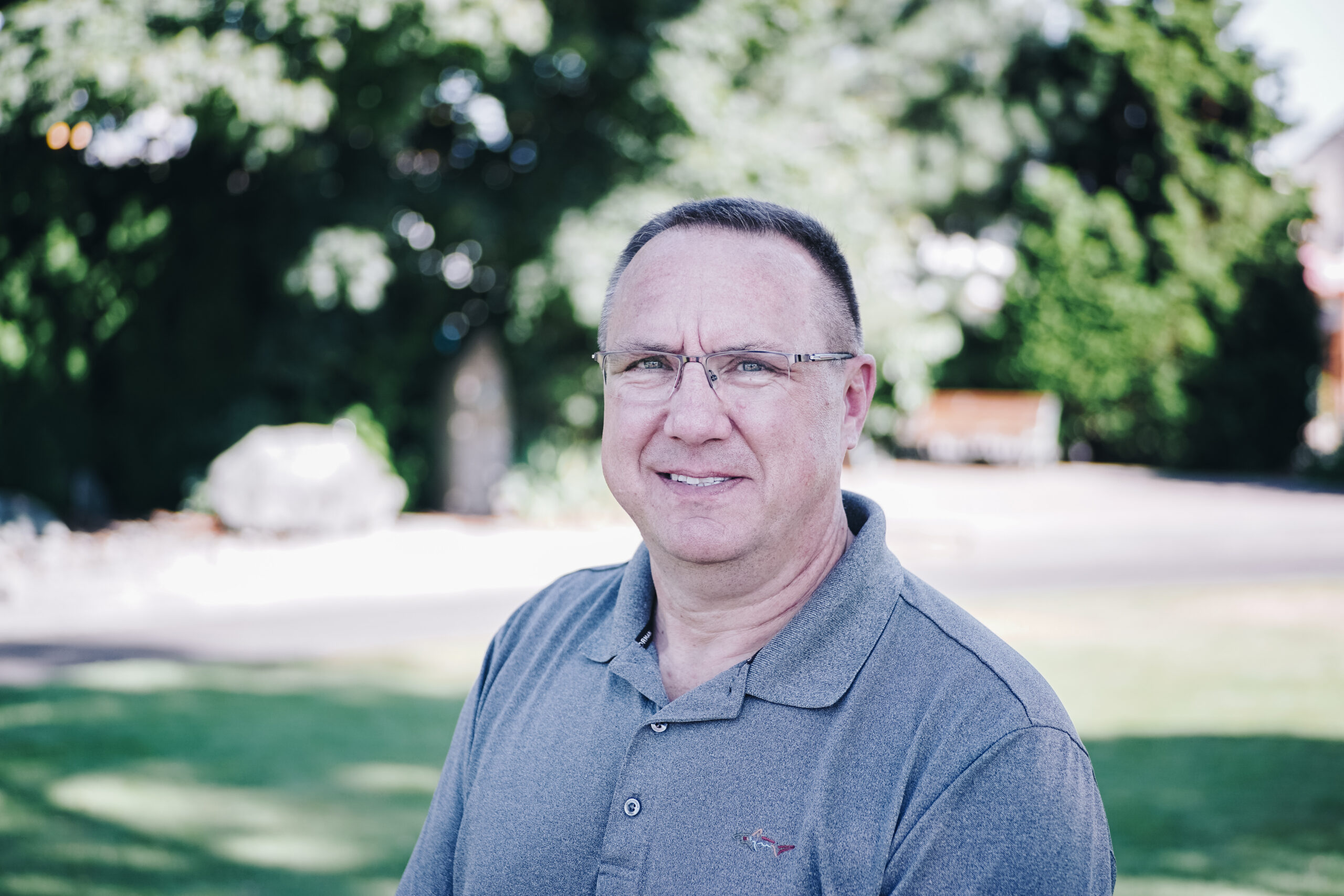 Middle-aged man with short hair and glasses wearing a grey polo shirt smiling outdoors.