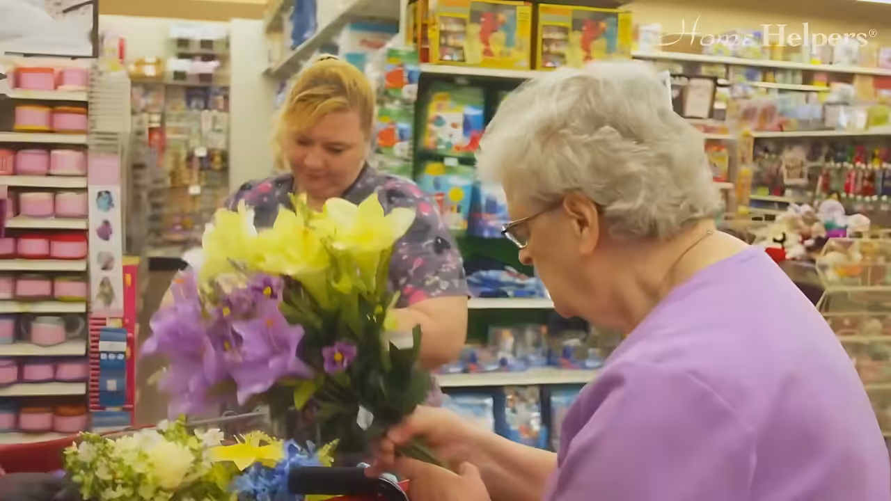 An elderly woman holding a bouquet of colorful flowers in a store, while a younger woman stands nearby assisting her. Shelves filled with various products are visible in the background.