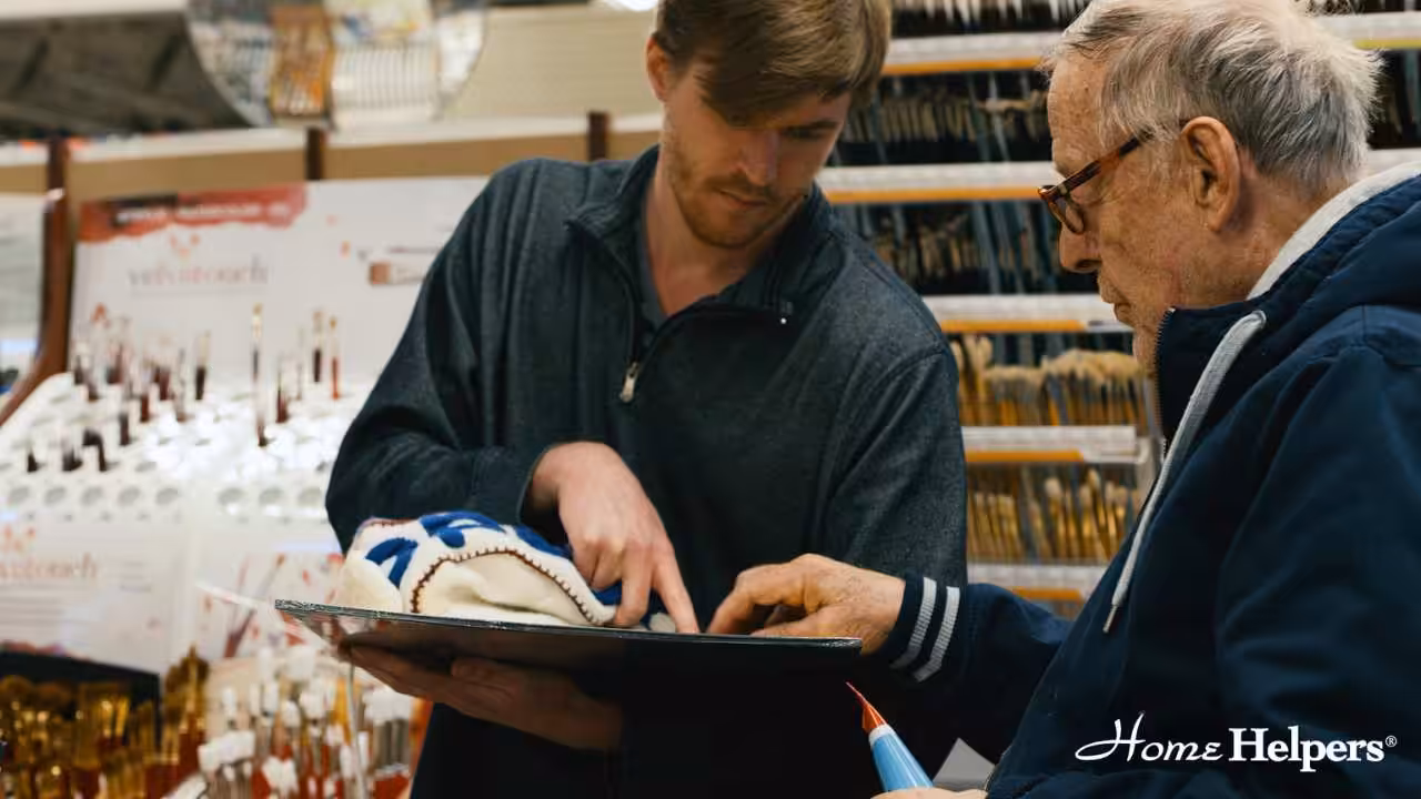 A younger man helps an older man look at information on a clipboard in a store aisle. The older man holds a shopping basket, and paintbrushes are displayed in the background. The Home Helpers logo appears in the corner.