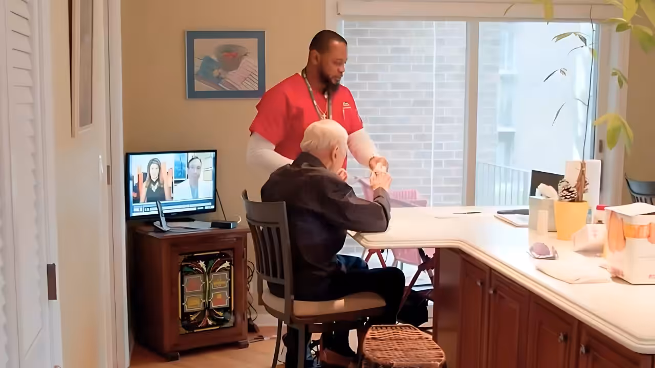 A caregiver in red scrubs assists an elderly man sitting at a kitchen counter. A TV with news anchors is on in the background, and sunlight streams through a sliding glass door.