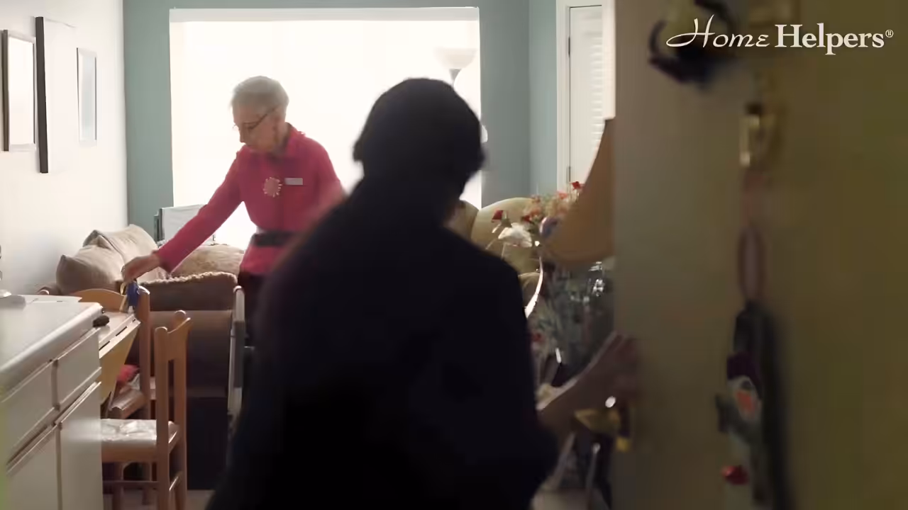A person enters a cozy, well-lit living room where an older woman with short gray hair, wearing a pink top, is standing with a walker. The Home Helpers logo appears in the top right corner.