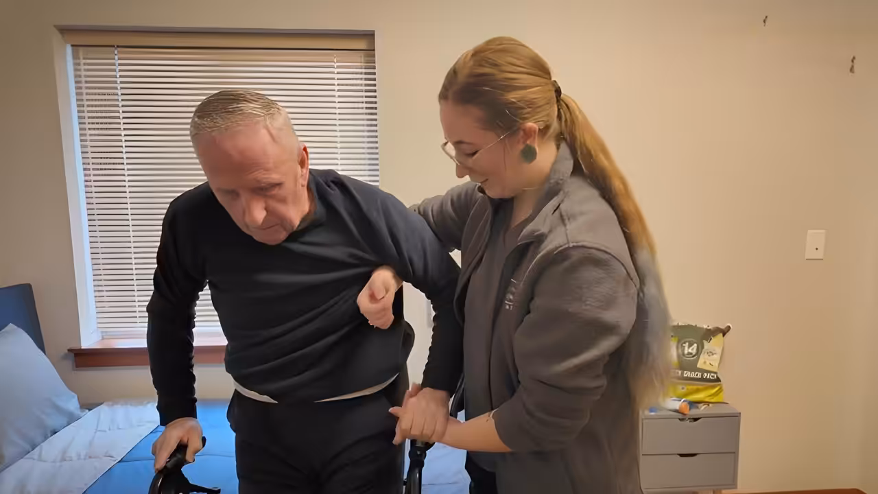A caregiver assists an older man with gray hair in standing up from a bed using a walker in a bedroom. The caregiver smiles while supporting the man by his arm and waist.