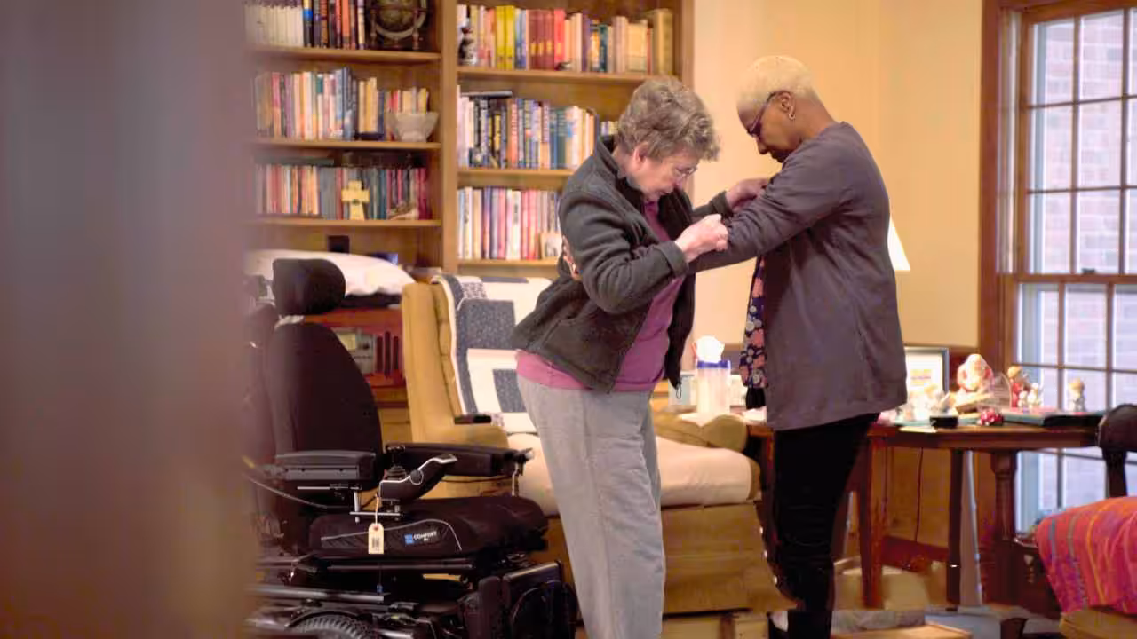 An older woman receives assistance from another woman while standing in a cozy room with bookshelves, a window, and a power wheelchair nearby.