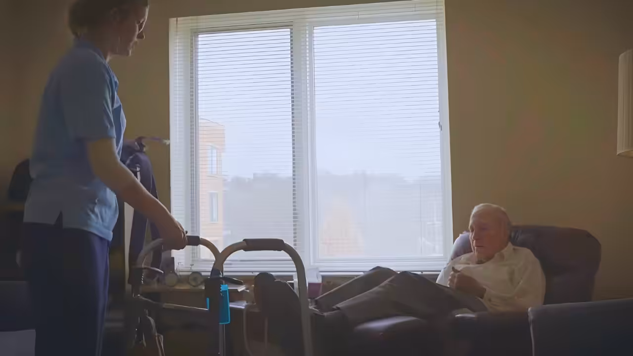 A caregiver stands beside a walker, assisting an elderly man who is sitting in a recliner by a window in a softly lit room.