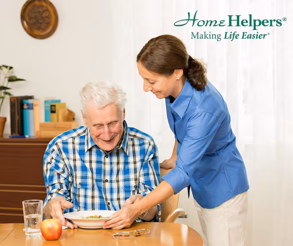A smiling elderly man sits at a table while a caregiver in a blue shirt serves him a meal. The table has an apple, a glass of water, and a book. Home Helpers - Making Life Easier is written in the corner.