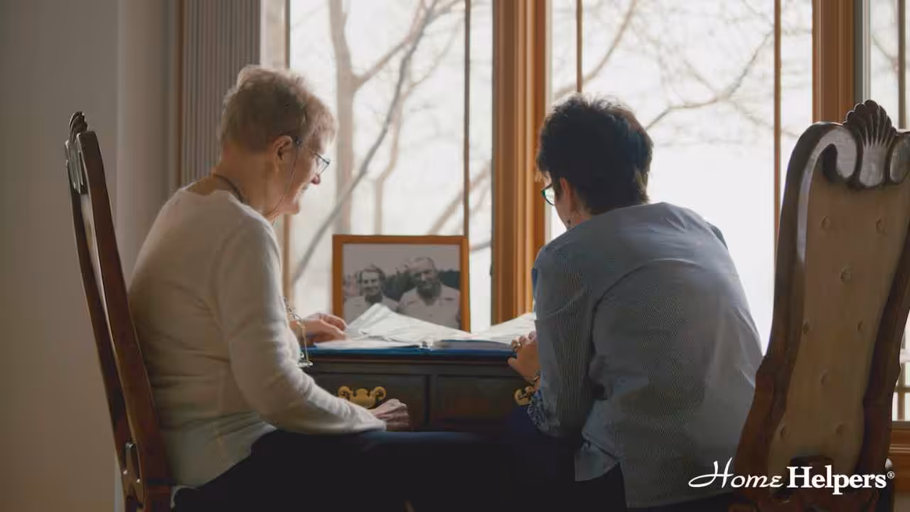 Two older adults sit at a desk by a window, looking at papers together. A framed photo of two people is on the desk. The logo Home Helpers appears in the bottom right corner.