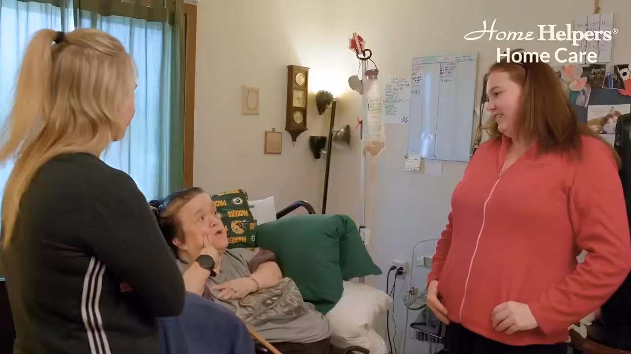 Two women stand and talk with a woman sitting in a bed in a cozy, well-lit room. The room has medical equipment, a whiteboard, and the Home Helpers Home Care logo in the upper right corner.