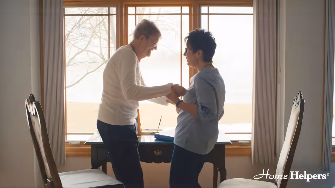 Two women stand smiling and holding hands in front of a window with a scenic outdoor view. They appear to be in a bright room with a wooden desk and chairs. The Home Helpers logo is visible in the bottom right corner.